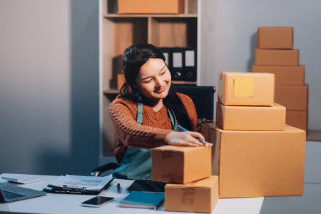 Startup SME small business entrepreneur of freelance Asian woman wearing apron using laptop and box to receive and review orders online to prepare to pack sell to customers, online sme business ideas.の写真素材