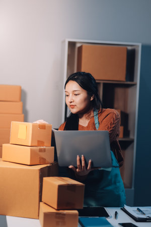 Startup SME small business entrepreneur of freelance Asian woman wearing apron using laptop and box to receive and review orders online to prepare to pack sell to customers, online sme business ideas.の写真素材