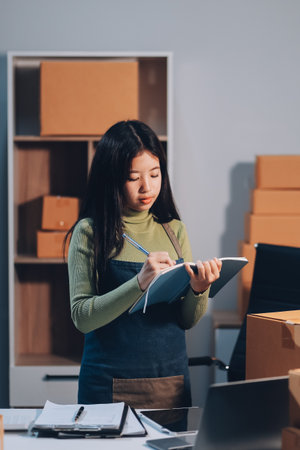 Startup SME small business entrepreneur of freelance Asian woman wearing apron using laptop and box to receive and review orders online to prepare to pack sell to customers, online sme business ideas.の写真素材