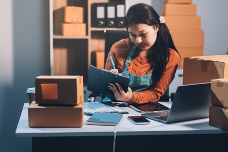 Startup SME small business entrepreneur of freelance Asian woman wearing apron using laptop and box to receive and review orders online to prepare to pack sell to customers, online sme business ideas.の写真素材
