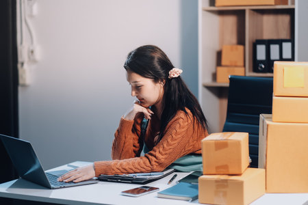 Startup SME small business entrepreneur of freelance Asian woman wearing apron using laptop and box to receive and review orders online to prepare to pack sell to customers, online sme business ideas.の写真素材