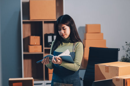Startup SME small business entrepreneur of freelance Asian woman wearing apron using laptop and box to receive and review orders online to prepare to pack sell to customers, online sme business ideas.の写真素材