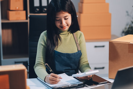 Startup SME small business entrepreneur of freelance Asian woman wearing apron using laptop and box to receive and review orders online to prepare to pack sell to customers, online sme business ideas.の写真素材