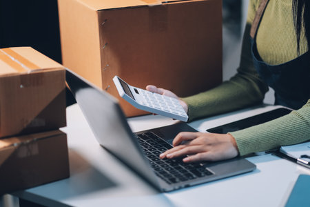 Startup SME small business entrepreneur of freelance Asian woman wearing apron using laptop and box to receive and review orders online to prepare to pack sell to customers, online sme business ideas.の写真素材