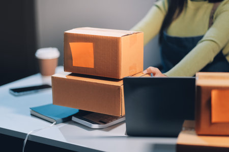 Startup SME small business entrepreneur of freelance Asian woman wearing apron using laptop and box to receive and review orders online to prepare to pack sell to customers, online sme business ideas.の写真素材