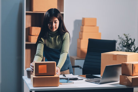 Startup SME small business entrepreneur of freelance Asian woman wearing apron using laptop and box to receive and review orders online to prepare to pack sell to customers, online sme business ideas.の写真素材