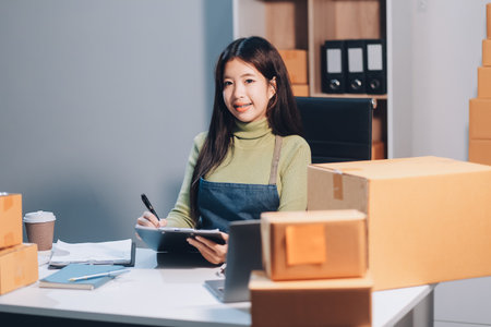 Startup SME small business entrepreneur of freelance Asian woman wearing apron using laptop and box to receive and review orders online to prepare to pack sell to customers, online sme business ideas.の写真素材