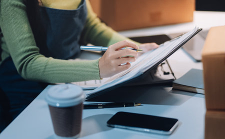 Startup SME small business entrepreneur of freelance Asian woman wearing apron using laptop and box to receive and review orders online to prepare to pack sell to customers, online sme business ideas.の写真素材