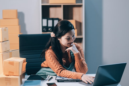 Startup SME small business entrepreneur of freelance Asian woman wearing apron using laptop and box to receive and review orders online to prepare to pack sell to customers, online sme business ideas.の写真素材