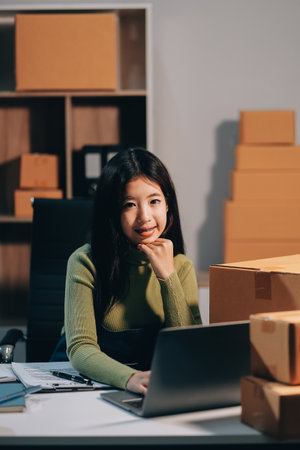 Startup SME small business entrepreneur of freelance Asian woman wearing apron using laptop and box to receive and review orders online to prepare to pack sell to customers, online sme business ideas.の写真素材