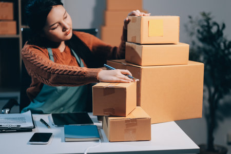Startup SME small business entrepreneur of freelance Asian woman wearing apron using laptop and box to receive and review orders online to prepare to pack sell to customers, online sme business ideas.の写真素材