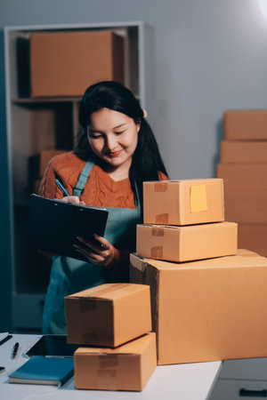 Startup SME small business entrepreneur of freelance Asian woman wearing apron using laptop and box to receive and review orders online to prepare to pack sell to customers, online sme business ideas.の写真素材