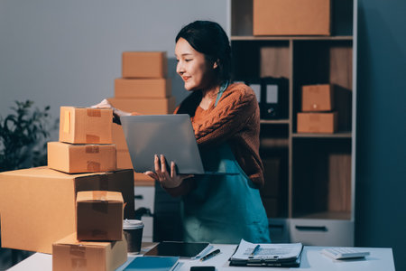 Startup SME small business entrepreneur of freelance Asian woman wearing apron using laptop and box to receive and review orders online to prepare to pack sell to customers, online sme business ideas.の写真素材