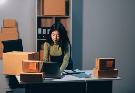 Startup SME small business entrepreneur of freelance Asian woman wearing apron using laptop and box to receive and review orders online to prepare to pack sell to customers, online sme business ideas.の写真素材