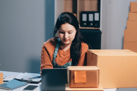 Startup SME small business entrepreneur of freelance Asian woman wearing apron using laptop and box to receive and review orders online to prepare to pack sell to customers, online sme business ideas.の写真素材