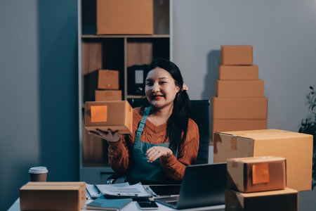 Startup SME small business entrepreneur of freelance Asian woman wearing apron using laptop and box to receive and review orders online to prepare to pack sell to customers, online sme business ideas.の写真素材