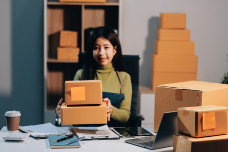 Startup SME small business entrepreneur of freelance Asian woman wearing apron using laptop and box to receive and review orders online to prepare to pack sell to customers, online sme business ideas.の写真素材