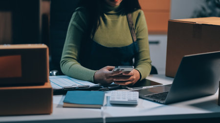 Startup SME small business entrepreneur of freelance Asian woman wearing apron using laptop and box to receive and review orders online to prepare to pack sell to customers, online sme business ideas.の写真素材