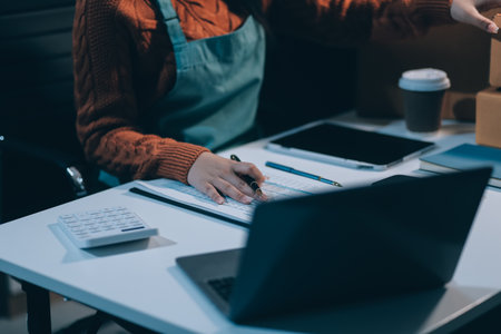 Startup SME small business entrepreneur of freelance Asian woman wearing apron using laptop and box to receive and review orders online to prepare to pack sell to customers, online sme business ideas.の写真素材