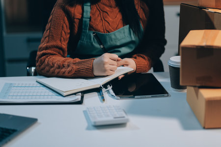 Startup SME small business entrepreneur of freelance Asian woman wearing apron using laptop and box to receive and review orders online to prepare to pack sell to customers, online sme business ideas.の写真素材