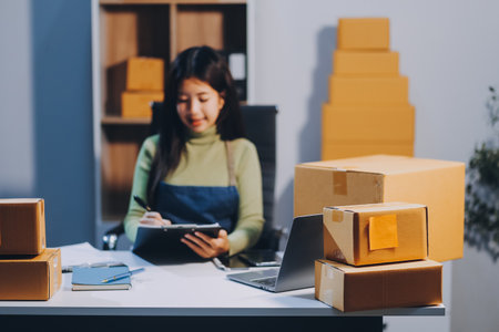 Startup SME small business entrepreneur of freelance Asian woman wearing apron using laptop and box to receive and review orders online to prepare to pack sell to customers, online sme business ideas.の写真素材