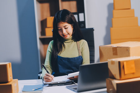 Startup SME small business entrepreneur of freelance Asian woman wearing apron using laptop and box to receive and review orders online to prepare to pack sell to customers, online sme business ideas.の写真素材