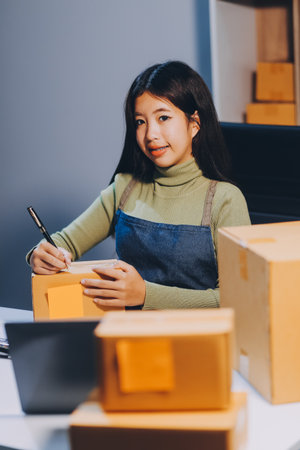 Startup SME small business entrepreneur of freelance Asian woman wearing apron using laptop and box to receive and review orders online to prepare to pack sell to customers, online sme business ideas.の写真素材