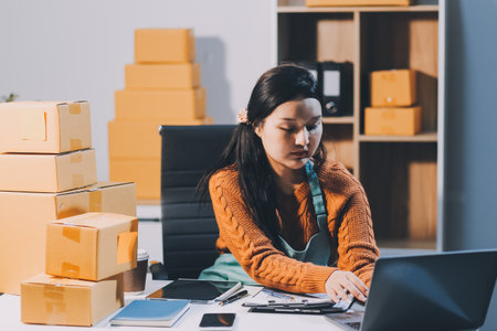 Startup SME small business entrepreneur of freelance Asian woman wearing apron using laptop and box to receive and review orders online to prepare to pack sell to customers, online sme business ideas.の写真素材