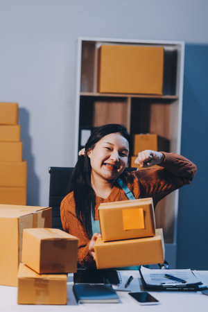 Startup SME small business entrepreneur of freelance Asian woman wearing apron using laptop and box to receive and review orders online to prepare to pack sell to customers, online sme business ideas.の写真素材