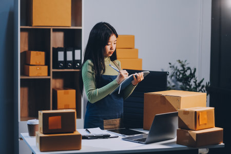 Startup SME small business entrepreneur of freelance Asian woman wearing apron using laptop and box to receive and review orders online to prepare to pack sell to customers, online sme business ideas.の写真素材