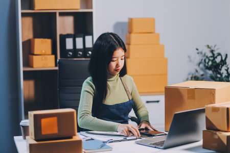 Startup SME small business entrepreneur of freelance Asian woman wearing apron using laptop and box to receive and review orders online to prepare to pack sell to customers, online sme business ideas.の写真素材