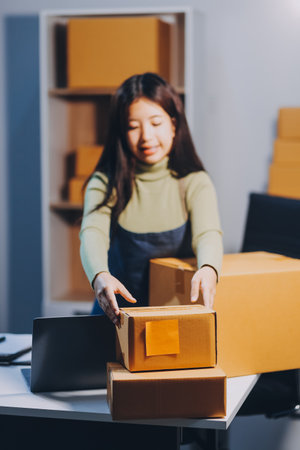 Startup SME small business entrepreneur of freelance Asian woman wearing apron using laptop and box to receive and review orders online to prepare to pack sell to customers, online sme business ideas.の写真素材