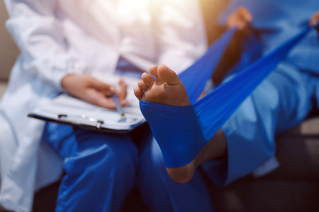 Confident physical therapist helps patient use resistance band stretching out his leg in clinic room.の写真素材