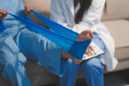 Confident physical therapist helps patient use resistance band stretching out his leg in clinic room.の写真素材
