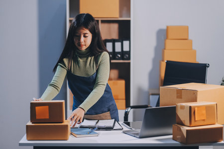 Startup SME small business entrepreneur of freelance Asian woman wearing apron using laptop and box to receive and review orders online to prepare to pack sell to customers, online sme business ideas.の写真素材