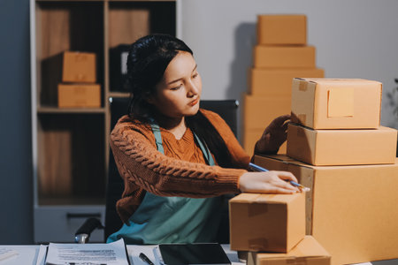 Startup SME small business entrepreneur of freelance Asian woman wearing apron using laptop and box to receive and review orders online to prepare to pack sell to customers, online sme business ideas.の写真素材