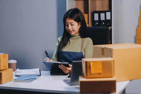 Startup SME small business entrepreneur of freelance Asian woman wearing apron using laptop and box to receive and review orders online to prepare to pack sell to customers, online sme business ideas.の写真素材