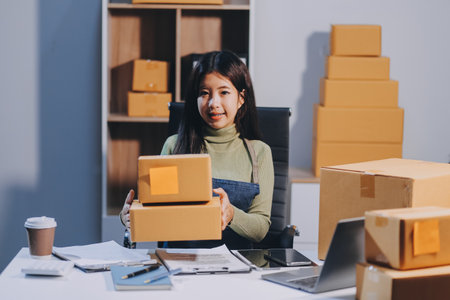 Startup SME small business entrepreneur of freelance Asian woman wearing apron using laptop and box to receive and review orders online to prepare to pack sell to customers, online sme business ideas.の写真素材