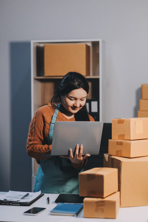 Startup SME small business entrepreneur of freelance Asian woman wearing apron using laptop and box to receive and review orders online to prepare to pack sell to customers, online sme business ideas.の写真素材
