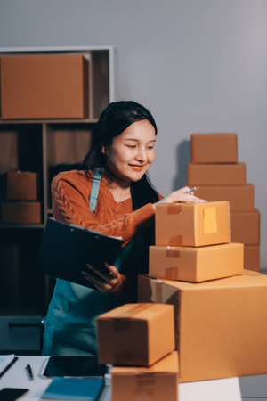 Startup SME small business entrepreneur of freelance Asian woman wearing apron using laptop and box to receive and review orders online to prepare to pack sell to customers, online sme business ideas.の写真素材
