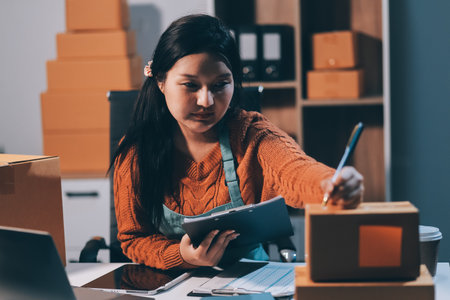 Startup SME small business entrepreneur of freelance Asian woman wearing apron using laptop and box to receive and review orders online to prepare to pack sell to customers, online sme business ideas.の写真素材