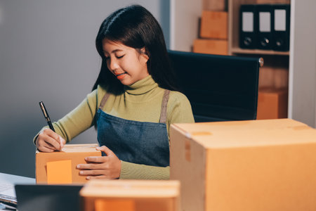 Startup SME small business entrepreneur of freelance Asian woman wearing apron using laptop and box to receive and review orders online to prepare to pack sell to customers, online sme business ideas.の写真素材