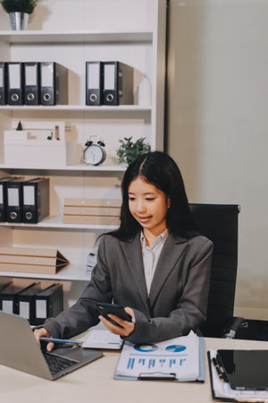 Young, smiling Asian businesswoman holding a jacket on her shoulder while using a mobile phone,...