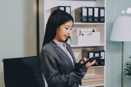 Young, smiling Asian businesswoman holding a jacket on her shoulder while using a mobile phone, standing confidently in a stylish, modern office environmentの写真素材