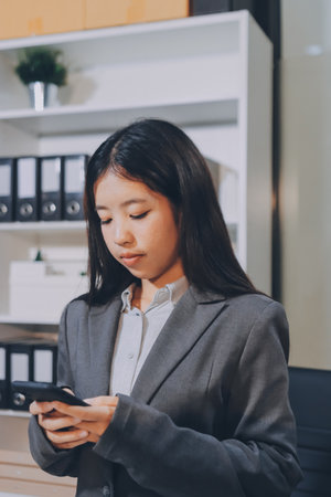 Young, smiling Asian businesswoman holding a jacket on her shoulder while using a mobile phone, standing confidently in a stylish, modern office environmentの写真素材