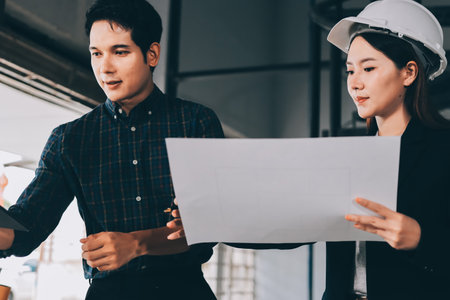 Construction manager and engineer dressed in orange work vests and hard helmets explore construction documentation on the building site near the steel framesの写真素材