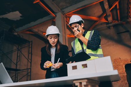 Construction manager and engineer dressed in orange work vests and hard helmets explore construction documentation on the building site near the steel framesの写真素材