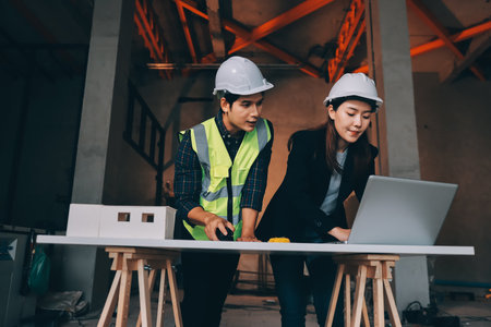 Construction manager and engineer dressed in orange work vests and hard helmets explore construction...