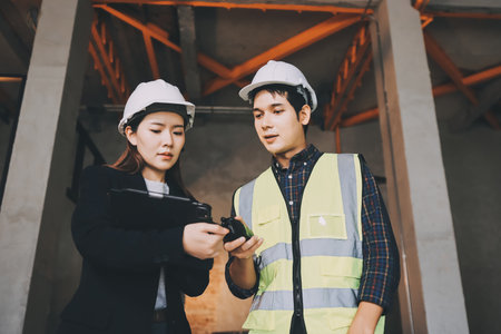 Construction manager and engineer dressed in orange work vests and hard helmets explore construction documentation on the building site near the steel framesの写真素材
