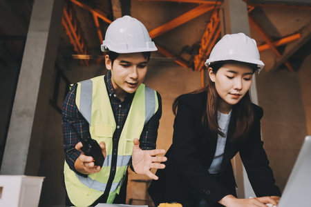 Construction manager and engineer dressed in orange work vests and hard helmets explore construction...